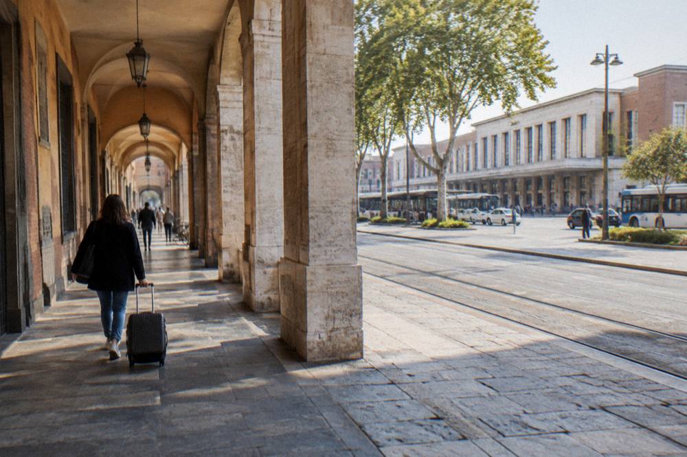 A person walking a small wheeled case along a narrow arcaded street in an Italian city