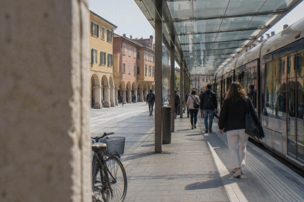 Modern railway station forecourt with a tram and passengers under a long glass canopy
