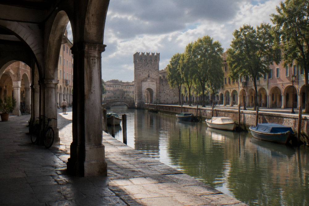 Stone city gate beside a canal with moored boats and a tree-lined embankment