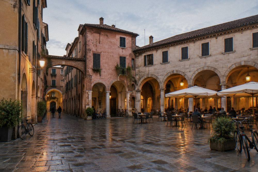 Arcaded buildings and cafe tables in an old Italian square lit by warm lamps in the evening