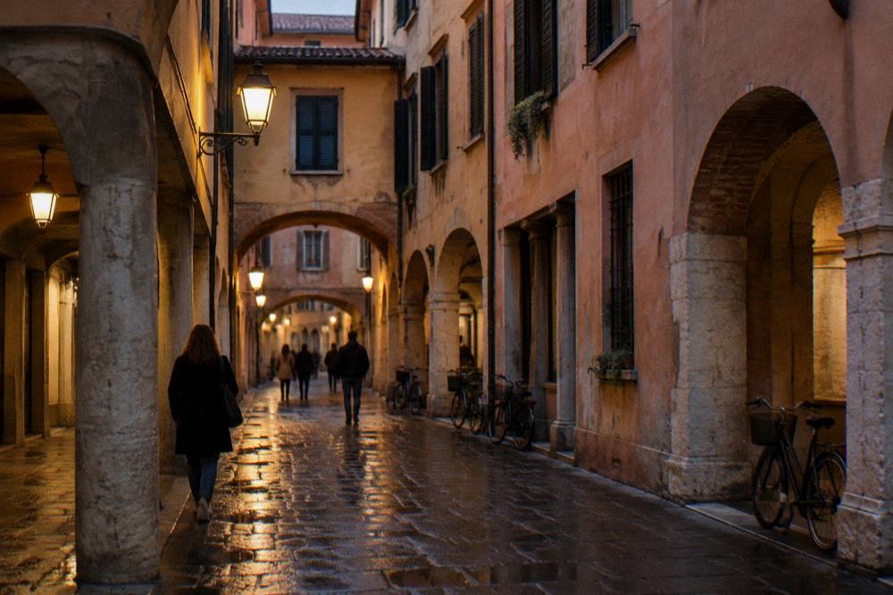 A lamplit pedestrian street in an old Italian city at night with people walking under arcades