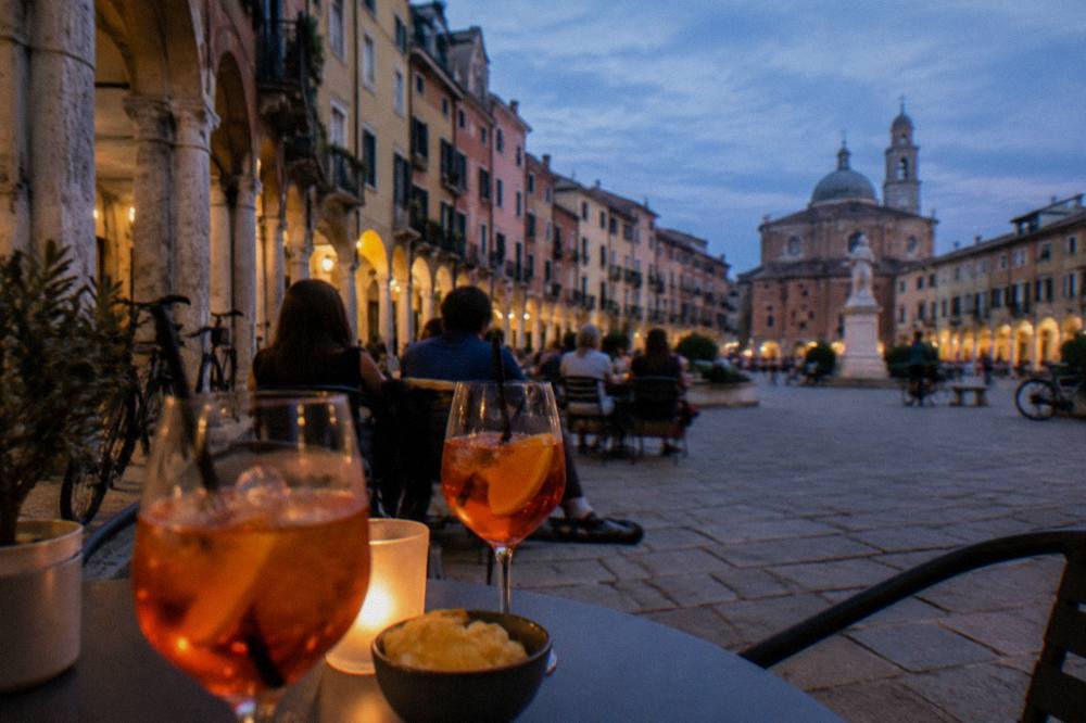 People seated at outdoor tables in a Padua square in the early evening with glasses of spritz on the table