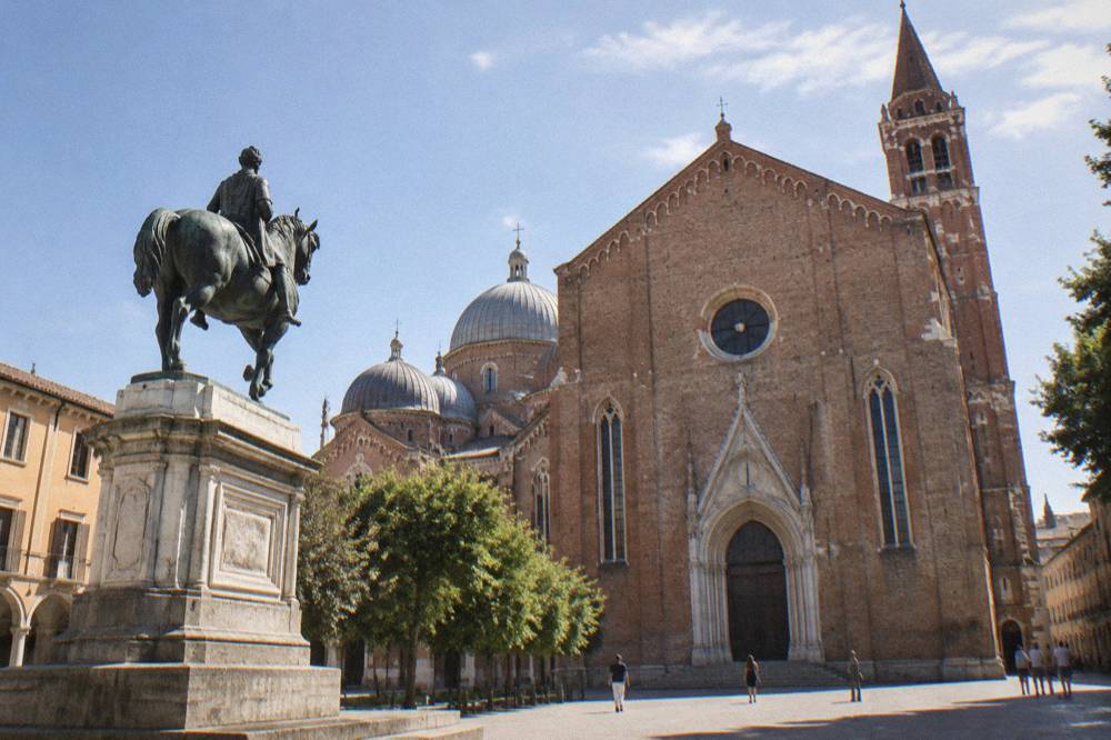 The brick gothic facade and domes of a large basilica seen from a square with an equestrian statue