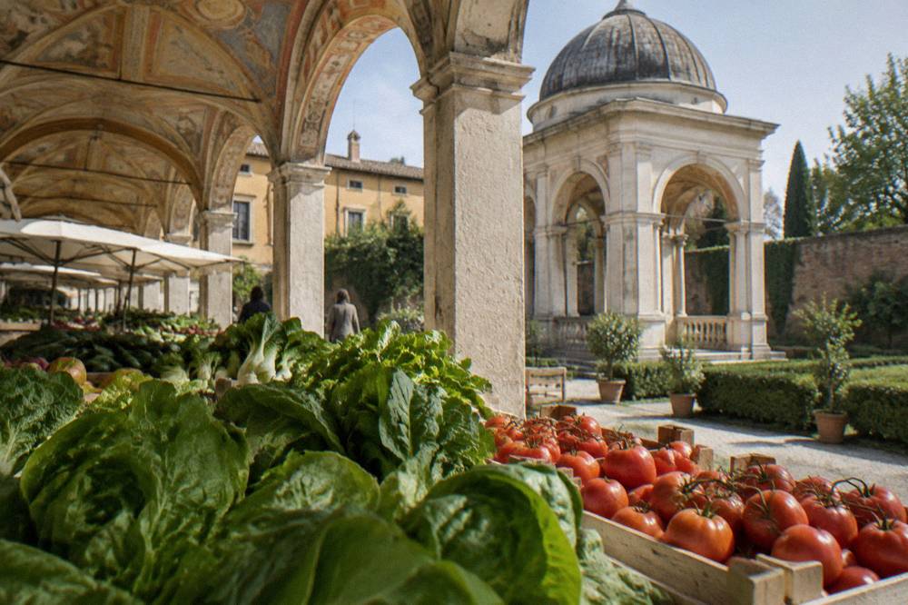 Stalls of leafy vegetables and crates of tomatoes beneath the painted facade of an Italian market loggia