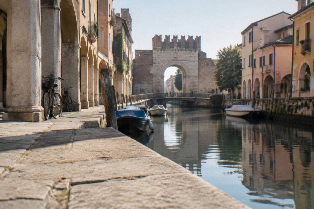 An old stone gate beside a calm canal with moored small boats in Padua