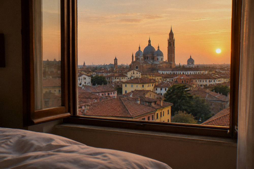 A hotel room window overlooking red rooftops and church domes of Padua at sunset