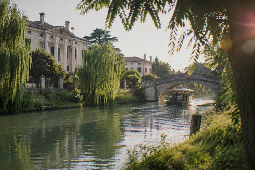 A flat-bottomed cruise boat passing under a stone bridge on a Veneto canal