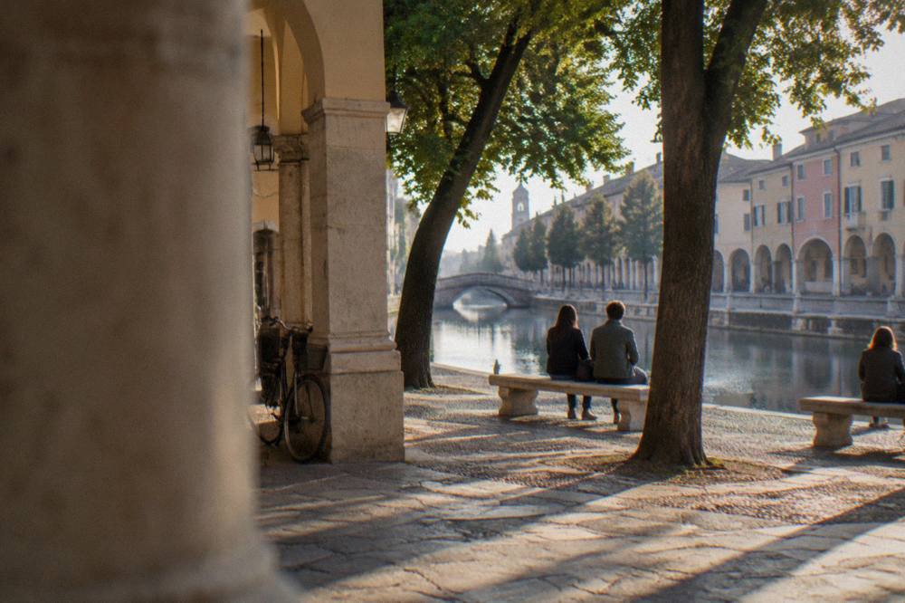 Visitors resting on benches under trees beside the canal ring of a large Padua square