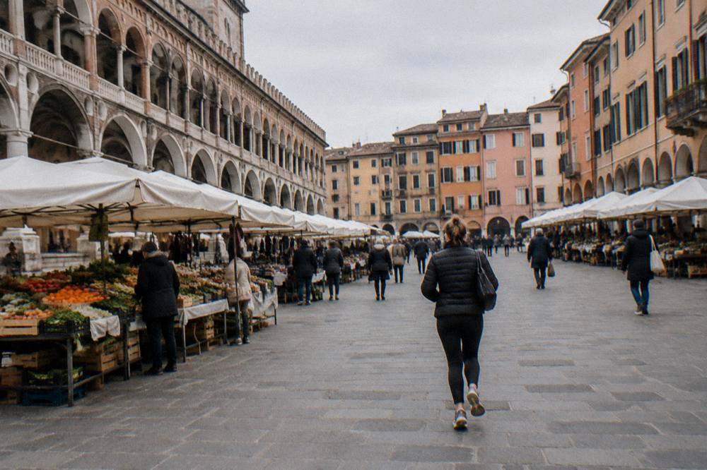 A wide Italian square at midday with market stalls, arcades and people walking